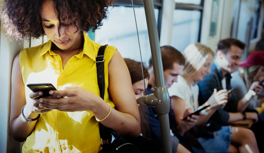 Black business woman in yellow shirt standing and scrolling on phone in a busy train